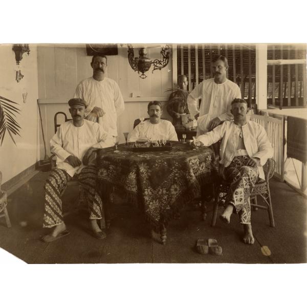 A photo of five International Drillers wearing white shirts, around a table with patterned tablecloth. There is a set of glasses on the table. There is a servant standing behind a plant against a white wall.