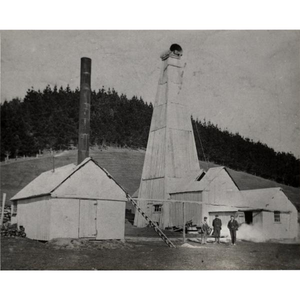 A photo of three men standing in front of an oil rig covered in wooden planks. There is a white building with a door at the front and a chimney coming out of the roof on the left. There is a line of trees in the background.