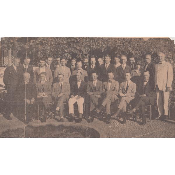 A group of men in two rows. One row is standing and the other is sitting in chairs. They are wearing suits and are in front of large bushes. 