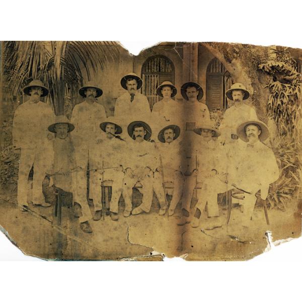 Twelve International Drillers outside of a building with a palm tree behind them. They are wearing white suits and pith helmets. Six men are standing and six are sitting on chairs.