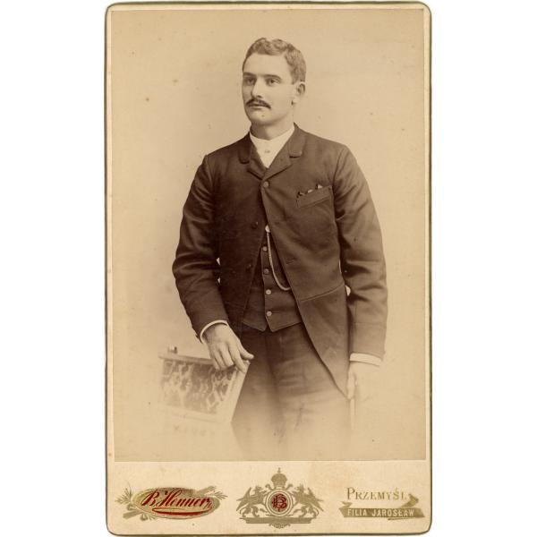 A photo of George Vansickle from the knees up. He is wearing a dark jacket, vest, and pants. His right arm is resting on the back of a chair.