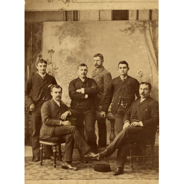 A photo of six men in front of a painted backdrop. They are wearing dark suits and two are sitting in chairs. There is a patterned rug underneath their feet.