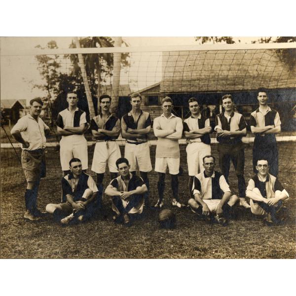 Twelve men wearing soccer uniforms in front of a net. Eight of the men are standing (seven with their arms folded) and four are sitting. There is a ball in the middle of the seated players.  