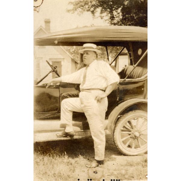 A photo of Fred Webb standing with his foot up on the step of a car. Webb is wearing a white shirt and pants, a tie, and a boat hat. The car has a canvas top and no windows. There is a building with a peaked roof in the background. 