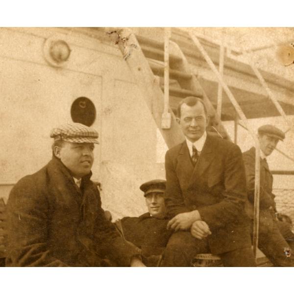 A photo of four men sitting at the base of a set of metal stairs on a boat deck. They are wearing dark jackets. 