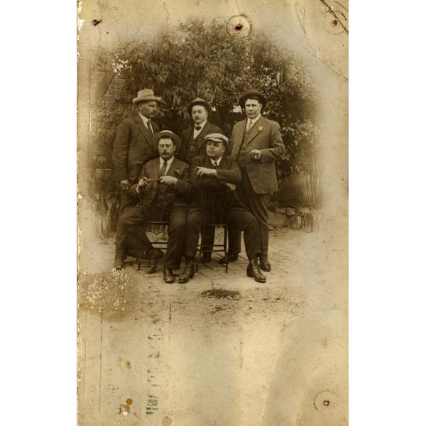 A photo of five International Drillers wearing dark suits and hats. Three are standing and two are sitting on chairs that are facing backwards. There is a tree behind them. 