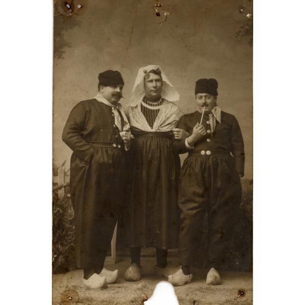 A photo of three men, wearing clogs. The man in the middle is wearing a skirt as well as a kerchief on his head. The men on either side are wearing high-waisted pants with two buttons and kerchiefs tied around their necks. They both have pipes.  