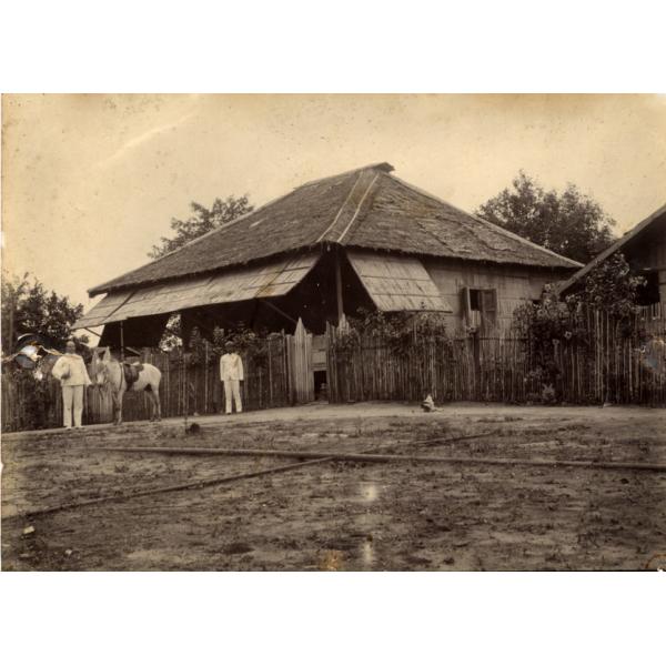 A photo of two men with a saddle donkey in front of a fence outside a thatched bungalow with the windows propped open by sticks. They are wearing white.  