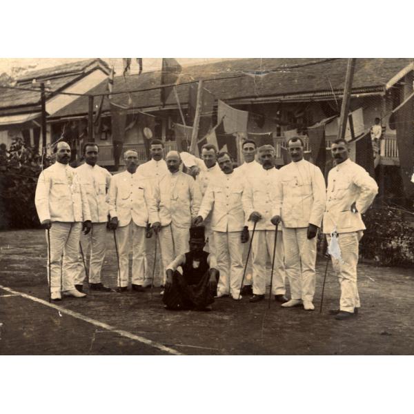 Une photo de onze foreurs internationaux portant des costumes blancs et tenant des cannes. Un homme vêtu de vêtements foncés et d'un chapeau est assis à l'avant. Derrière, il y a une rangée de bâtiments avec des drapeaux triangulaires accrochés entre eux. 