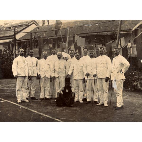 A photo of eleven International Drillers wearing white suits and holding canes. There is a man wearing dark clothes and a hat sitting in front. Behind, there is a row of buildings with triangular flags hanging between them. 