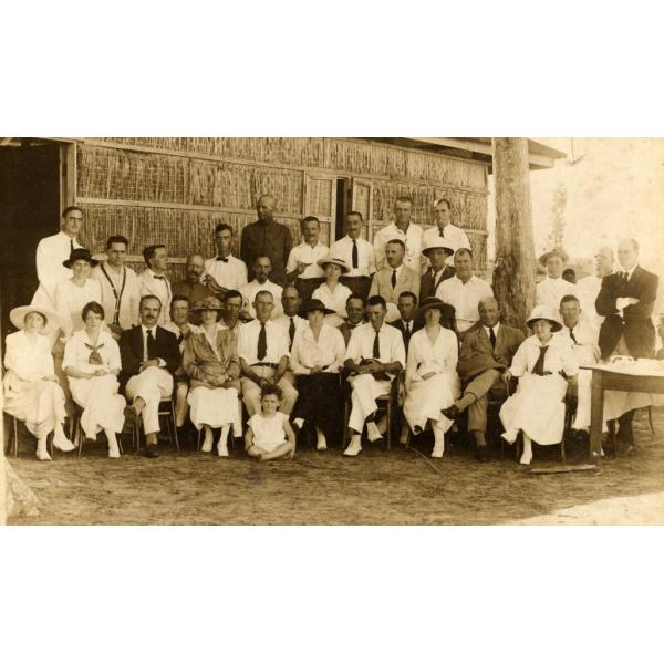 A photo of a group of men and women, with one child, in front of a thatched building. Most of the people are wearing white shirts. 