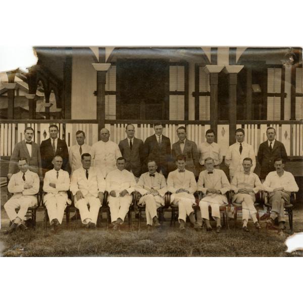 A photo of nineteen men in two rows. They are in front of a building with a deck and white railings. The back row is standing and the front row is sitting in chairs. Most of the men are wearing white.  