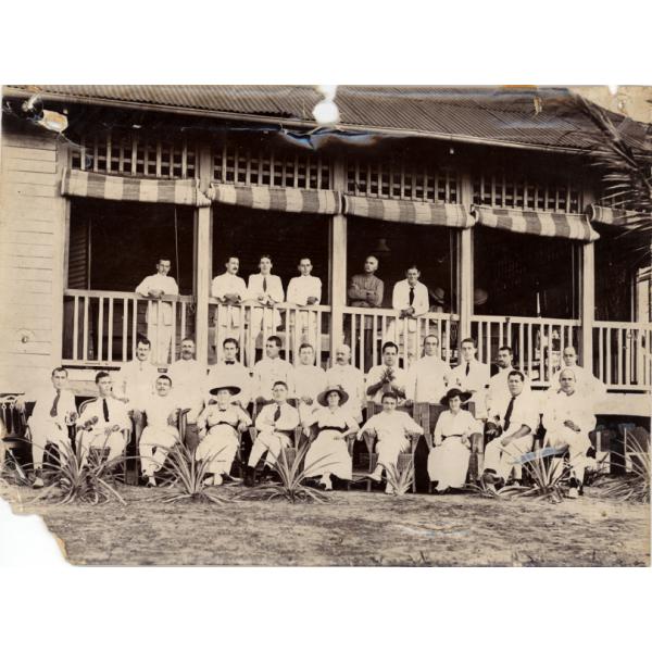 A photo of a group of men and three women, all wearing white. The top row stands on the deck of a building, against a railing. The middle row stands on the ground. The front row is seated in chairs, with spiky plants in front of them.  