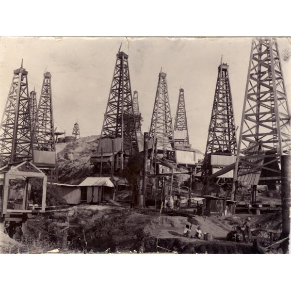 Une photo de dix tours de forage pour le pétrole rapprochées les unes des autres sur une colline. Certaines sont munies de réservoirs de stockage cylindriques en acier. Il y a une chaudière à vapeur, avec de grandes cheminées, au centre. 