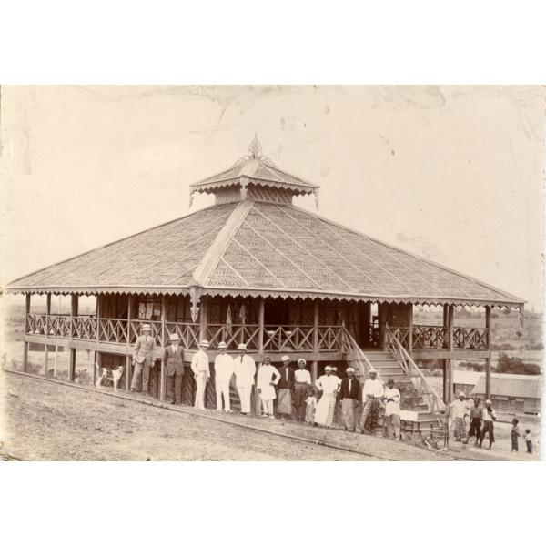 A photo of a group of men standing outside a wooden building with an ornate roof. There is a deck with a railing around the second floor with a set of stairs running up to it.