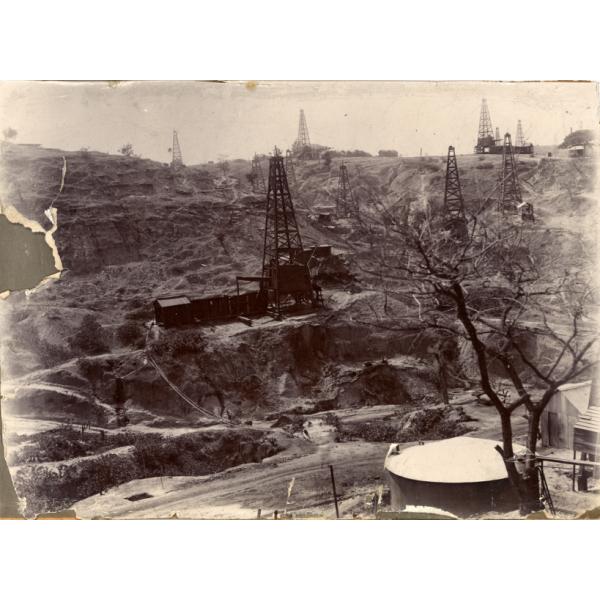 A photo of a dirt field on a hill. There are a dozen oil rigs dotting the hill with pipes running down to a storage tank in the bottom right corner.