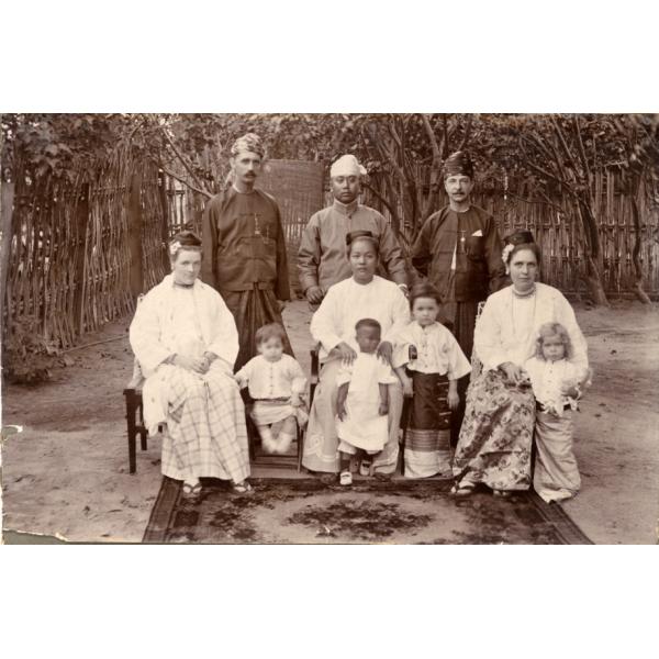 Three men stand behind three sitting women and four children. They are wearing simple shirts and skirts made from a large piece of fabric. There are trees and a wooden fence behind them, and a patterned rug at their feet.