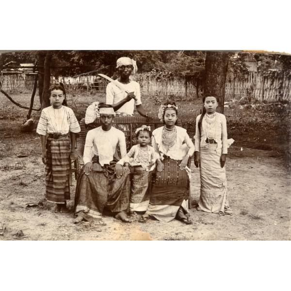 A photo of six Burmese people in traditional clothing. Three are sitting on a wicker chair, and there is a girls standing on either side. A man stands behind holding a machete.