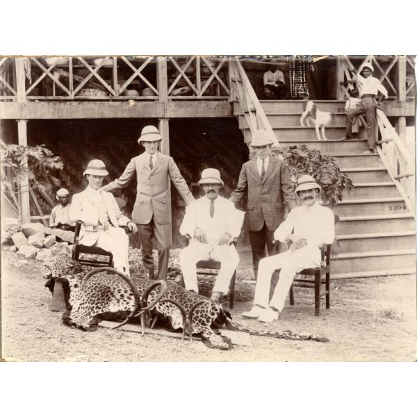 A photo of five men wearing pith helmets, two standing and three sitting in chairs, behind a large cat skin and two sets of animal horns. There are two people and a dog on a set of stairs leading up to a building behind the men.