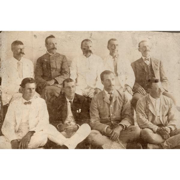 A photo of nine men. Five are sitting on a bench and four are sitting cross-legged on the floor. They are wearing a mixture of light and dark jackets.