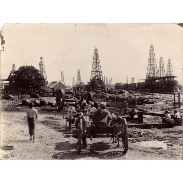 A photo of a group of men with oil derricks in the background. Some men are sitting on carts while others stand around a pile of casing. There is a set of large wagon wheels in the middle of the photo. 