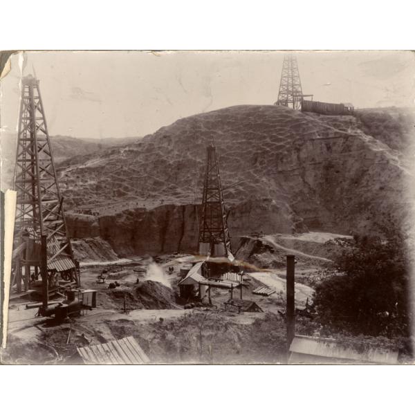 A photo of two metal oil rigs and a third rig up on a hill in behind. There are more hills in the background.