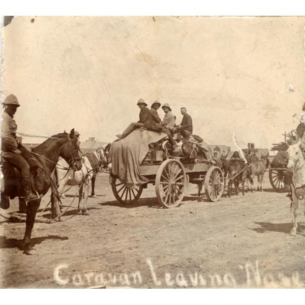 A photo of horse-drawn carts packed with goods. There are men riding on one of the carts and other men on horseback.