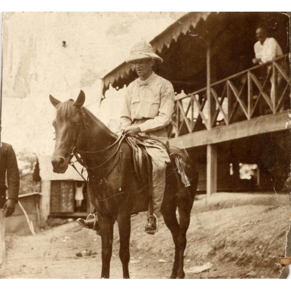 A photo of an International Driller wearing white clothing and a pith helmet, sitting on a horse. He is in front of a building that has been raised on stilts.