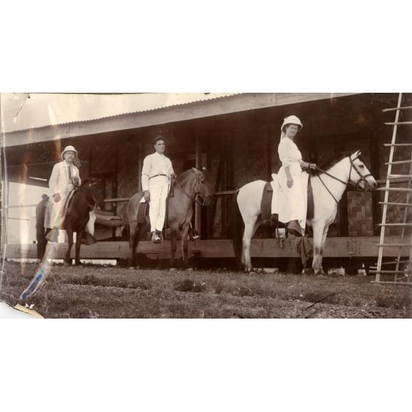 Deux hommes et une femme à cheval devant un bâtiment avec une terrasse surélevée. Les hommes montent des chevaux foncés et portent des chemises et des pantalons blancs. La femme est vêtue d'une robe blanche et d'un chapeau. Elle monte un cheval blanc. 