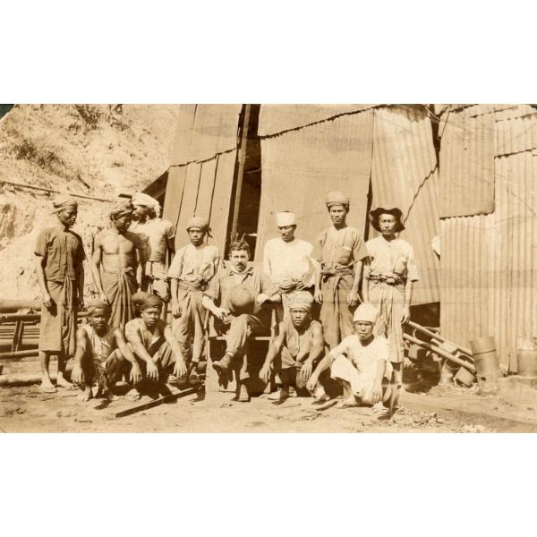 An International Driller sitting cross-legged on a chair in the middle of a group of workers. They are in front of a rig covered in sheet metal.