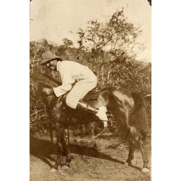 Une photo d'un homme accroupi sur un petit cheval foncé. Il porte un costume blanc, un pantalon, des chaussures et un chapeau plus foncé. Il y a des arbres en arrière-plan.  