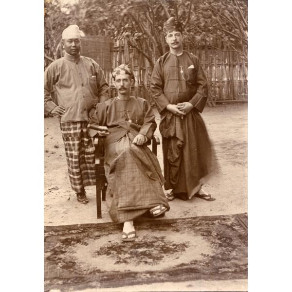 Un homme de la région et deux foreurs internationaux devant une clôture et des arbres, avec un tapis à leurs pieds. Ils portent des vestes amples et de longs morceaux de tissu noués comme une jupe ainsi que des tongs.