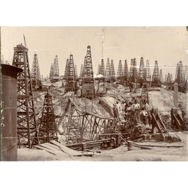 A field full of four-poled oil derricks. There is a group of people standing on a wooden footbridge that leads across a valley in the field. Casing, steam boilers, and cylindrical storage tanks are visible next to many of the derricks.