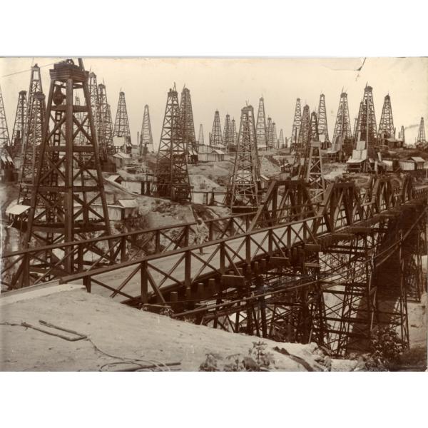 A field full of four-poled oil derricks behind a bridge spanning a low point in the field. There are buildings behind the first row of derricks.