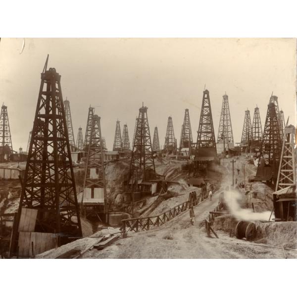 Close to thirty oil derricks with four poles each. There is a bridge leading across a low point in the oil field and several men are walking along the road. There are buildings behind the front row of derricks and a large groups of people are gathered near a pile of casing.