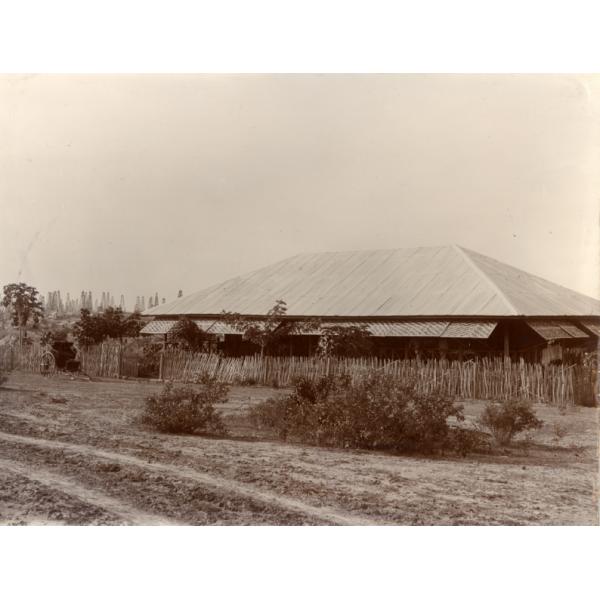 A bungalow with thatched window covers. It is surrounded by a fence made of sticks. There is a cart parked by the fence and a cluster of shrubs in front of it. There is a group of oil derricks in the background.