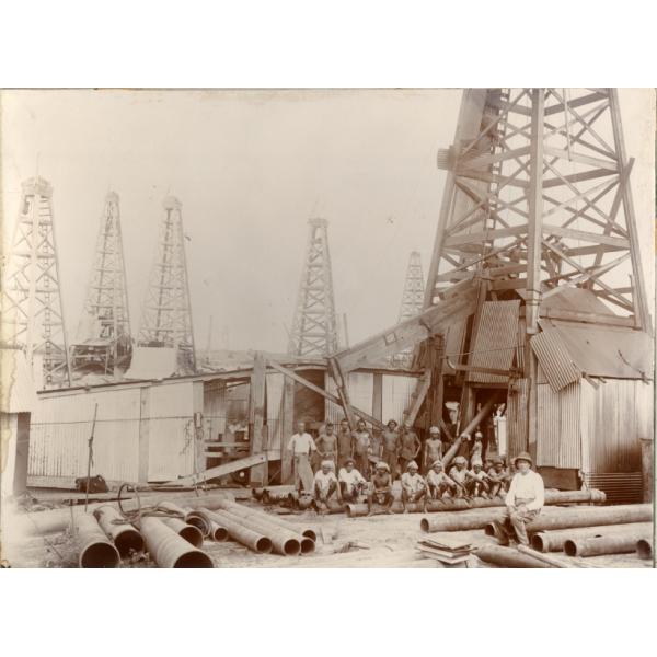 An oil crew in front of an oil rig. There are five oil rigs in the background and a pile of casing at the front of the photo. An International Driller is sitting on some of the casing.