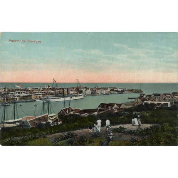 A postcard showing the entrance to a harbour.. There are boats docked along the edge of the light blue water. A group of people is standing on a hill looking at the boats. There are buildings very close to the edge of the water.  