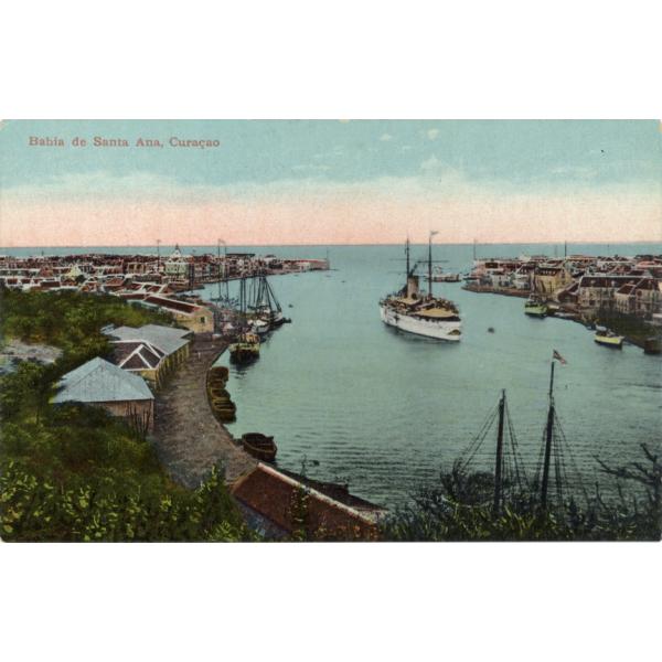 Une carte postale représentant une petite baie avec de l'eau bleu clair. Il y a un bateau à deux mâts qui vogue sur l'eau et un certain nombre de bateaux amarrés le long des quais. Des bâtiments se trouvent tout près du bord de l'eau. La carte postale indique « Baie de Santa Ana, Curaçao ». 