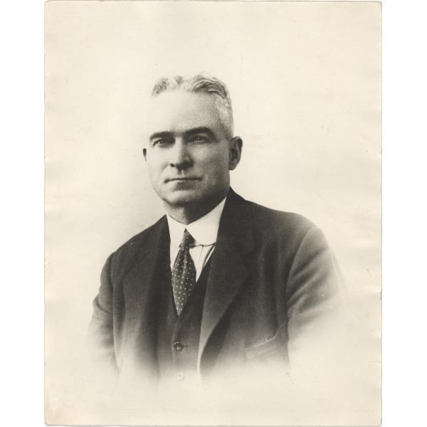 A photo of John Growder from the chest up. He is wearing a dark jacket and vest with a white shirt and dotted tie. He is clean-shaven with grey hair. 