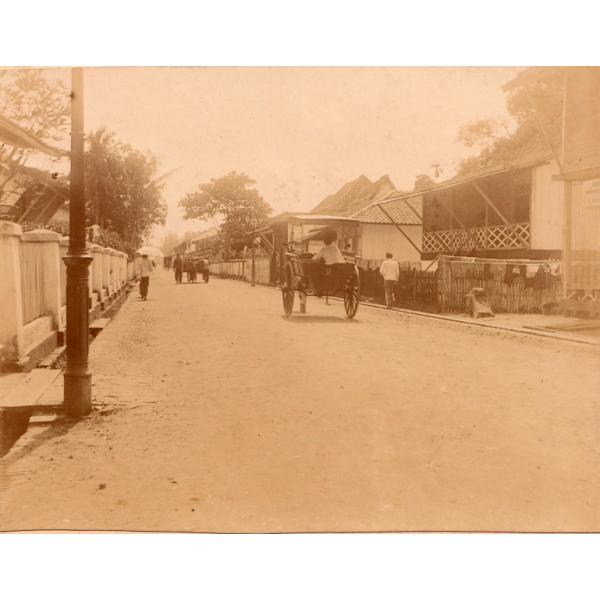  Un chemin de terre avec des bâtiments et des clôtures de chaque côté. Des gens marchent dans la rue et on peut aussi y voir une charrette couverte. 