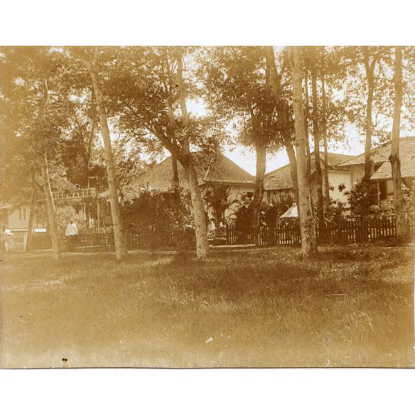 A man standing off to the left. There are buildings in the background with a fence and trees in front. 