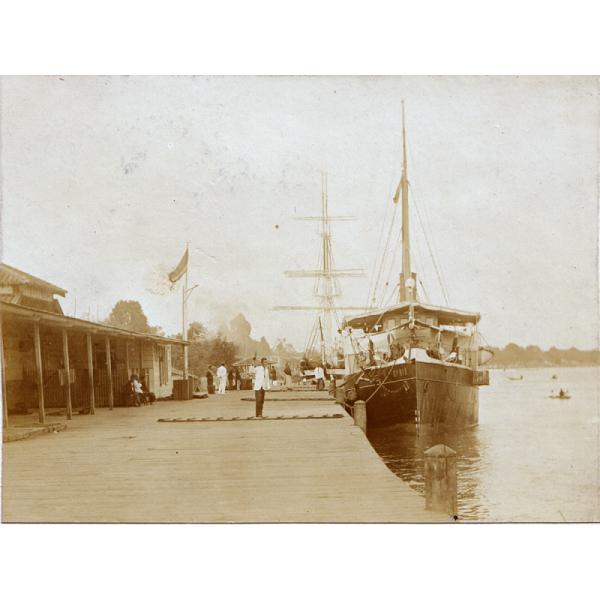 A ship docked alongside a wooden dock. There is another ship in behind and a building to the left. People are sitting and standing, looking at the ships. 