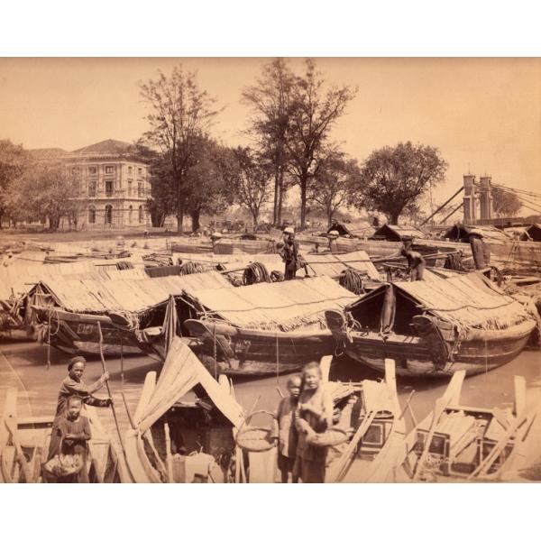 Photo d'un groupe de bateaux le long de la rive, avec des couvertures faites de roseaux. Il y a un grand bâtiment avec des fenêtres et des portes voûtées à l'arrière-plan, avec un groupe d'arbres. 