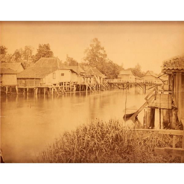 A photo of a group of buildings on stilts with thatched roofs. They are sitting directly above water and there is a bridge between the two sides of the river towards the back. There are trees in the background and reeds in the foreground.  