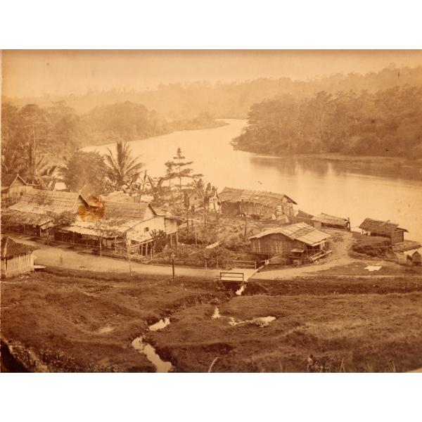 A photo of a group of buildings beside a curving river. There is a small stream running down a hill into the river. The road between the buildings has a small bridge over the stream. There are trees in the background. 