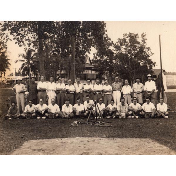 A baseball team. Half the men are standing and half are sitting. Most are wearing white t-shirts. They are in front of a group of trees and a building. There are three baseball bats set up like a tripod in front of them with a ball resting on top. Some other equipment is also in front of them. 