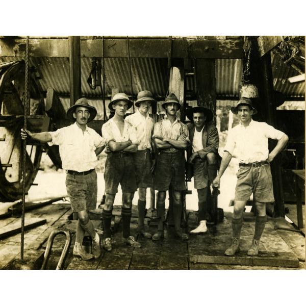 A photo of six International Drillers standing inside an oil rig with metal siding. They are wearing shorts, high socks, and white t-shirts.