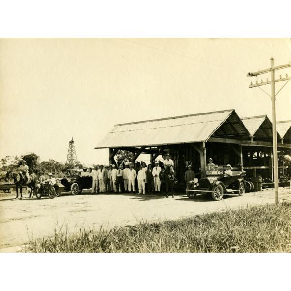 A group of people standing in front of an open wooden structure with a metal roof that has three peaks. There is a car and horse on either side of the group.