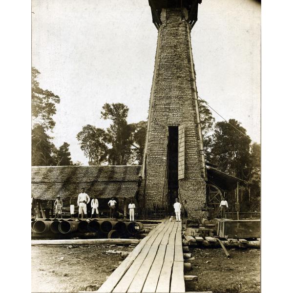 A photo of W.B.C. Isbister in a white suit and his dog standing on a plank path in front of a thatched oil rig. His crew stands to the left on a pile of casing.